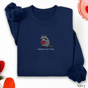 Può includere: Felpa blu navy con un procione ricamato che tiene un cuore rosso. Il testo "ROMANTIC BUT FERAL" è sotto il procione. La felpa ha maniche lunghe e girocollo.