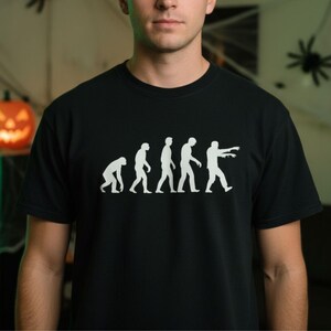 May include: Black t-shirt featuring a white graphic of the evolution of man, progressing into a zombie. The design shows a series of figures, starting hunched over and ending with a zombie reaching forward. The shirt is a crew neck and made of a soft material.
