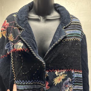 May include: A dark blue jacket with floral patchwork and striped accents. The jacket features a notched collar and is embellished with decorative stitching in white, red, and yellow. The jacket is displayed on a black mannequin.