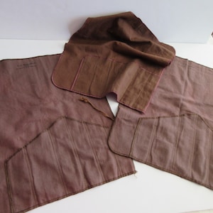 May include: A set of brown fabric tool rolls, featuring multiple pockets and a flap closure. The fabric has a slightly textured appearance, with darker brown stitching and accents. The rolls are designed to hold tools.