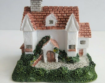 Może przedstawiać: Miniaturowy biały domek z czerwonym dachem z dachówek, ceglanym kominem i małym gankiem z drewnianymi drzwiami. Domek otoczony jest zielenią i kamienną ścieżką.