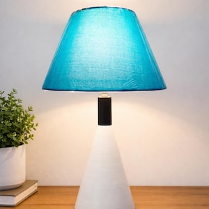 Puede incluir: Una lámpara de mesa moderna con una pantalla azul turquesa y una base cónica blanca. La lámpara tiene un cuello cilíndrico negro que conecta la pantalla a la base. La lámpara está sobre una superficie de madera.