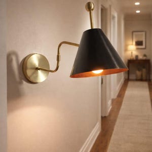 May include: A wall-mounted sconce with a black conical shade and a gold-colored interior. The sconce has a brass-colored arm and a circular base. The light is on, illuminating the interior of the shade and the surrounding wall.