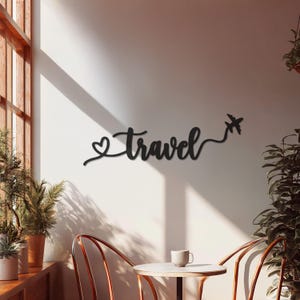May include: Black metal wall art spelling out the word "Travel" in a cursive font, with a heart symbol before the word and an airplane icon at the end. The art is mounted on a white wall.