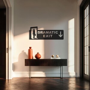 May include: A black metal sign with the words "DRAMATIC EXIT" and an arrow, above a dark wood console table. The table holds two orange vases and two small decorative spheres. The sign features a silhouette of a person.