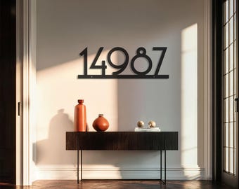May include: Black metal house number sign displaying the numbers 14987. The numbers are mounted on a horizontal bar. The sign is mounted on a white wall above a dark wood console table. Decorative orange bottles and spheres are on the table.