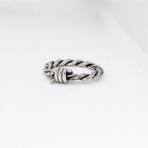 Pu&ograve; includere: Un anello color argento con un design a corda intrecciata. L'anello presenta una serie di elementi cilindrici impilati nella parte anteriore, aggiungendo un dettaglio unico. L'anello &egrave; posto su uno sfondo bianco.