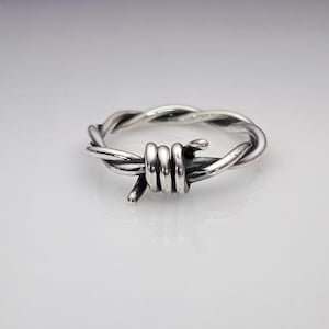 May include: A silver-colored ring designed to resemble barbed wire. The ring features twisted metal strands and sharp, pointed barbs. The design is a unique and edgy piece of jewelry.