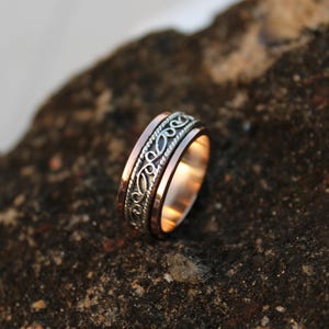 May include: A close-up of a two-tone ring with a copper band and a silver filigree design. The ring is set against a dark, textured background, highlighting its intricate details and warm tones. The ring's design features a repeating pattern.