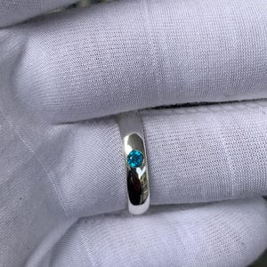 May include: A polished silver-toned band ring featuring a single, small, round, bright blue gemstone set flush into the centre of the metal surface. The ring has a smooth, reflective finish and a simple, clean design with a circular stone setting.