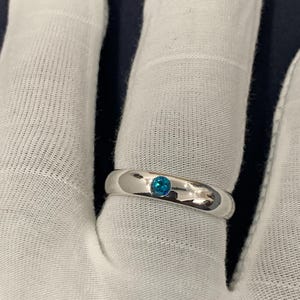 May include: A polished silver-tone band ring featuring a single, small, round, bright blue gemstone set flush into the centre of the metal surface. The ring has a smooth, rounded profile and a reflective finish, showcasing a clean and simple design.