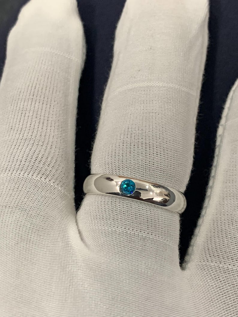 May include: A polished silver-tone band ring featuring a single, small, round, bright blue gemstone set flush into the centre of the metal surface. The ring has a smooth, rounded profile and a reflective finish, showcasing a clean and simple design.