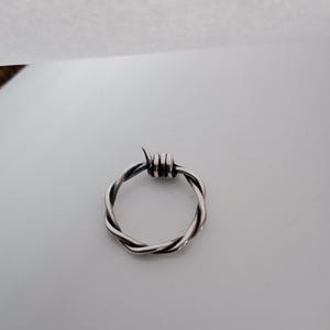 Pu&ograve; includere: Un anello color argento con un design a filo spinato. L'anello &egrave; circolare, con fili di metallo attorcigliati che formano la fascia e piccole punte nella parte superiore. L'anello &egrave; posto su uno sfondo bianco.