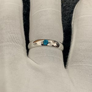 May include: A polished silver-tone band ring featuring a single, small, round, bright blue stone set flush into the centre of the metal surface. The ring has a smooth, rounded profile and a reflective finish, showcasing a minimalist design.