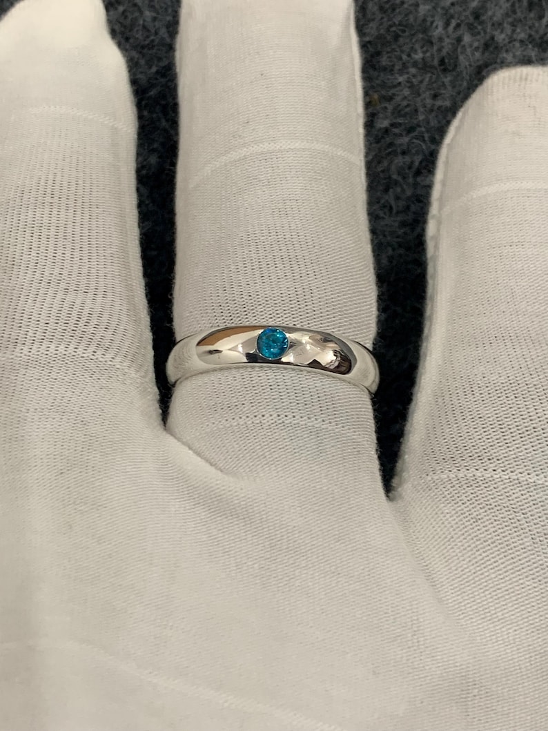 May include: A polished silver-tone band ring featuring a single, small, round, bright blue stone set flush into the centre of the metal surface. The ring has a smooth, rounded profile and a reflective finish, showcasing a minimalist design.