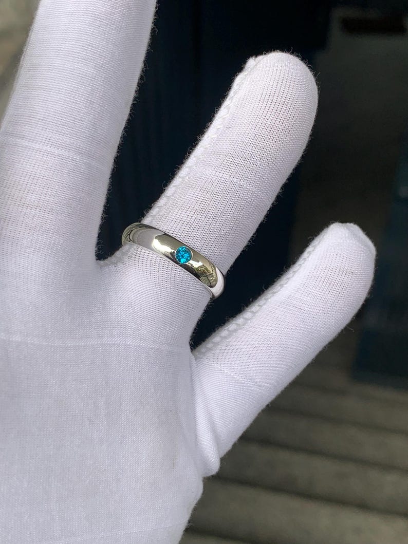 May include: A polished silver-toned band ring featuring a single, small, round, bright blue gemstone set flush into the centre of the metal surface. The ring has a smooth, reflective finish and a simple, rounded profile design.