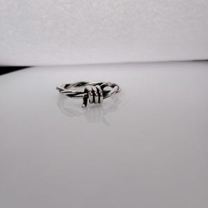 Pu&ograve; includere: Un anello color argento con un design a filo spinato. L'anello &egrave; realizzato in metallo lucido e presenta una rappresentazione dettagliata di filo spinato avvolto attorno alla fascia. L'anello &egrave; esposto su una superficie riflettente.