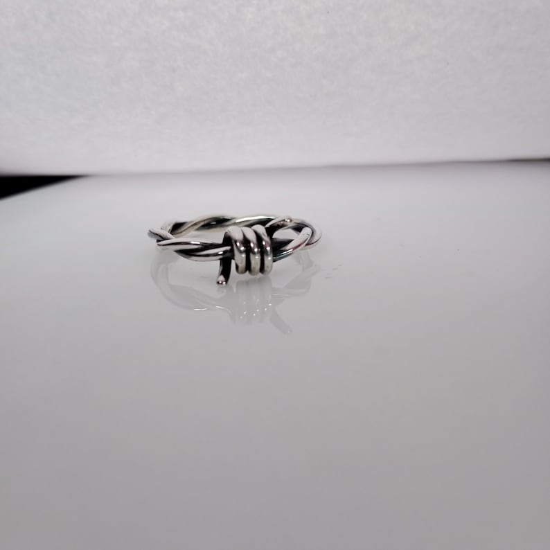 Pu&ograve; includere: Un anello color argento con un design a filo spinato. L'anello &egrave; realizzato in metallo lucido e presenta una rappresentazione dettagliata di filo spinato avvolto attorno alla fascia. L'anello &egrave; esposto su una superficie riflettente.