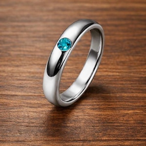 Puede incluir: Un anillo plateado con un acabado pulido, con una pequeña gema redonda de color turquesa. El anillo se encuentra sobre un fondo de madera cálida, resaltando su diseño elegante y el color vibrante de la piedra.