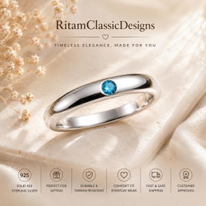 May include: A polished silver band ring featuring a single round, bright blue gemstone set in the centre. The text RitamClassicDesigns is visible at the top, with additional icons and text regarding 925 sterling silver at the bottom.