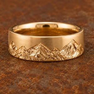 May include: A gold band ring featuring a textured, raised mountain range design around the exterior. The ring has a brushed, matte finish on the upper portion and a polished, reflective finish on the jagged, detailed mountain peaks and valleys.
