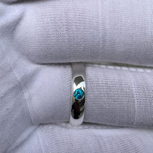 Puede incluir: Un anillo de plata pulida con una sola gema redonda de color turquesa brillante. La superficie pulida del anillo refleja la luz. El anillo se encuentra sobre un fondo de tela blanca.