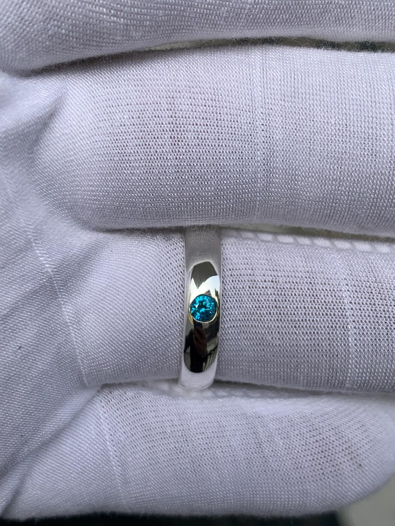 May include: A polished silver band ring featuring a single, round, brilliant-cut bright blue gemstone set flush into the centre of the metal surface. The ring has a smooth, reflective finish and a simple, clean design with no additional ornamentation.