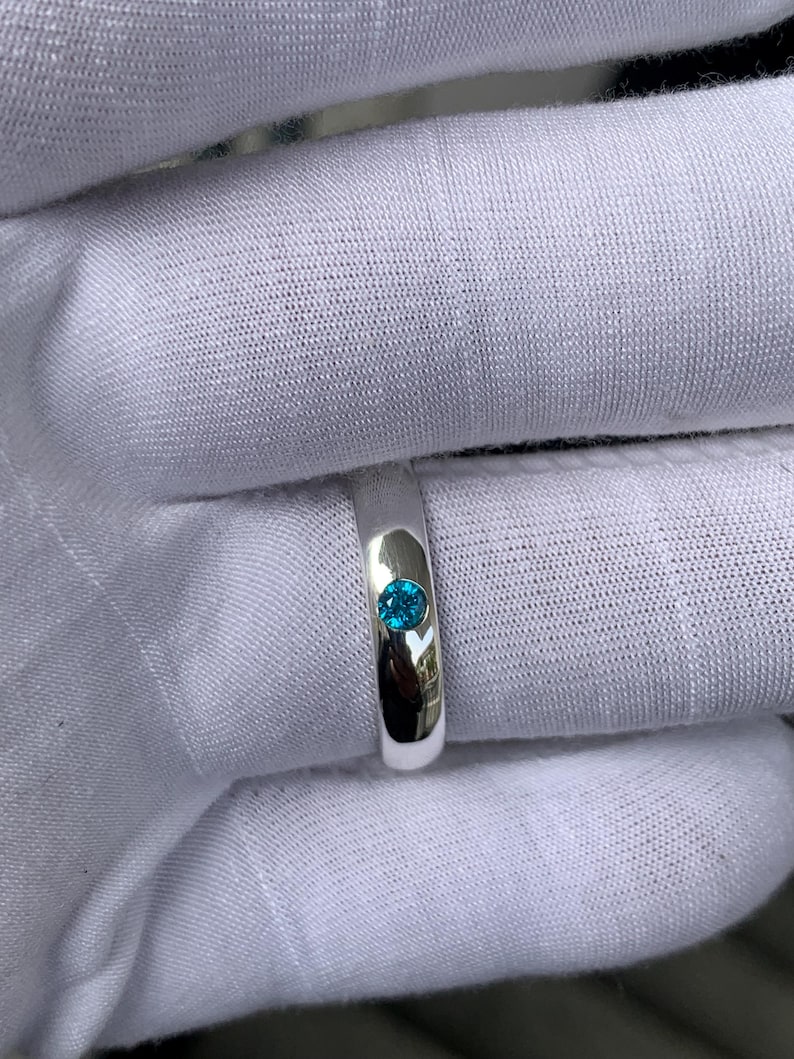 May include: A polished silver-toned band ring featuring a single, small, round, bright blue gemstone set flush into the centre of the metal surface. The ring has a smooth, reflective finish and a simple, clean design with a circular stone setting.