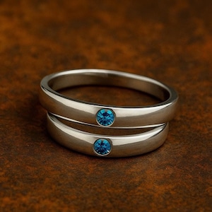 Puede incluir: Dos anillos de plata apilados, cada uno con una piedra preciosa azul redonda. Los anillos tienen un acabado liso y pulido y están sobre un fondo marrón. Las piedras preciosas son de un tono azul vibrante.