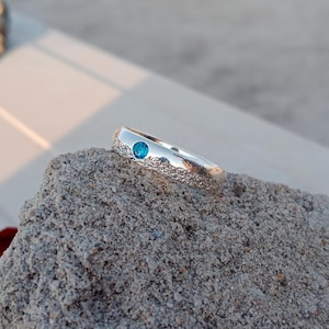 Może przedstawiać: Srebrny pierścionek z teksturowanym paskiem i małym, okrągłym, jasno niebieskim kamieniem szlachetnym. Pierścionek jest umieszczony na chropowatej, szarej kamiennej powierzchni, z rozmytym tłem.