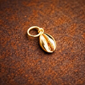 May include: A gold-toned metal charm shaped like a small cowrie shell, featuring a smooth, polished surface and a central ridged opening. The charm is attached to a circular jump ring, designed for use as a pendant or jewelry component.