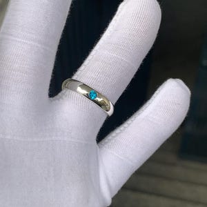 May include: A polished silver-toned band ring featuring a single, small, round, bright blue gemstone set flush into the centre of the metal surface. The ring has a smooth, reflective finish and a simple, rounded profile design.