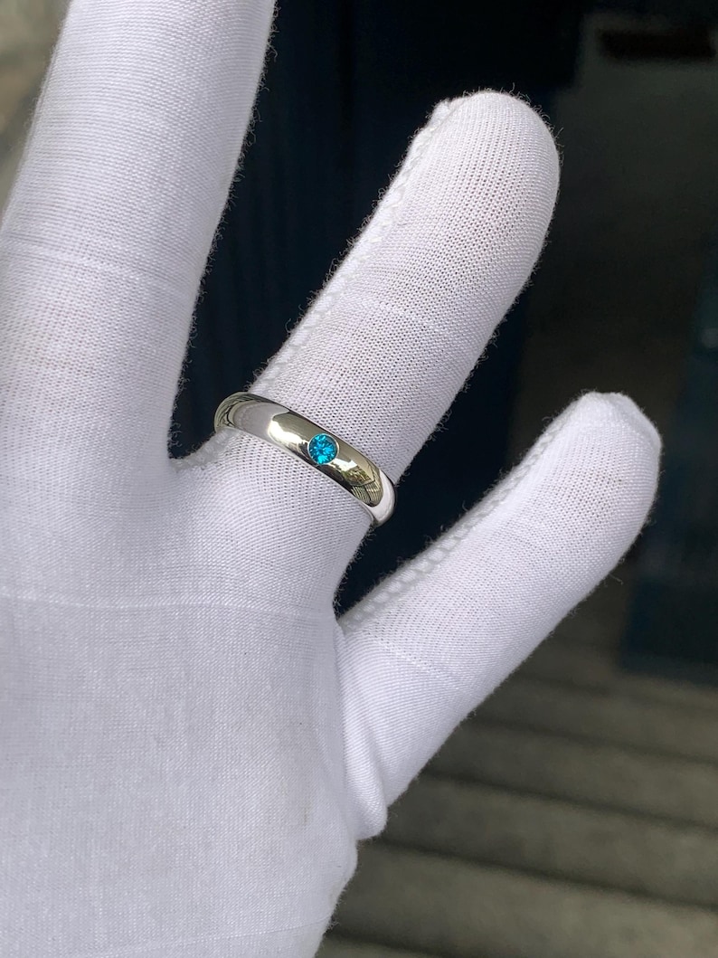 May include: A polished silver-toned band ring featuring a single, small, round, bright blue gemstone set flush into the centre of the metal surface. The ring has a smooth, reflective finish and a simple, rounded profile design.