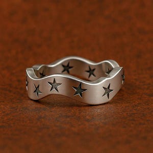 May include: A silver ring with a wavy design. The ring has cut-out star shapes around the band. The ring is shown on a brown surface.
