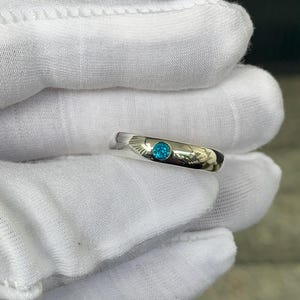 May include: A polished silver-toned metal band ring featuring a single, round, brilliant-cut bright blue gemstone set flush into the centre of the band. The ring has a smooth, reflective surface and a simple, clean design with a circular stone setting.