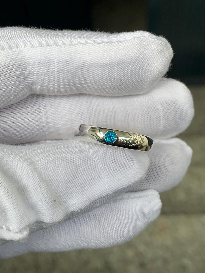 May include: A polished silver-toned metal band ring featuring a single, round, brilliant-cut bright blue gemstone set flush into the centre of the band. The ring has a smooth, reflective surface and a simple, clean design with a circular stone setting.