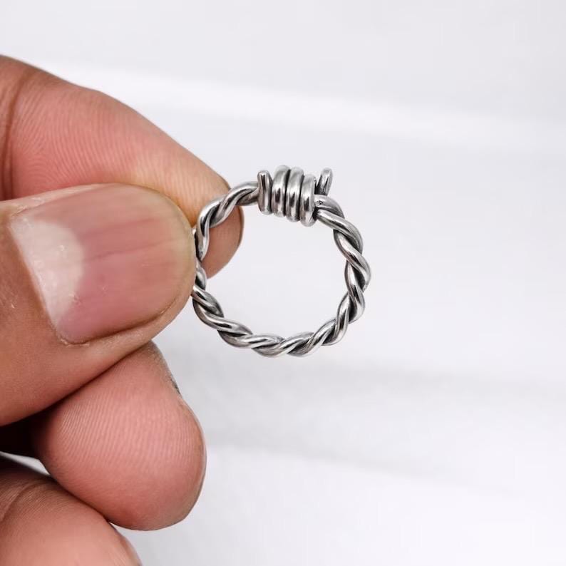 Pu&ograve; includere: Un anello color argento con un design a filo spinato. L'anello presenta una fascia intrecciata e una piccola sezione di filo avvolto, creando un'estetica unica e audace. L'anello &egrave; tenuto su uno sfondo bianco.