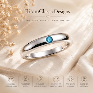May include: A polished silver band ring featuring a single round, bright blue gemstone set in the centre. The text RitamClassicDesigns is visible at the top, with additional icons and text regarding 925 sterling silver at the bottom.