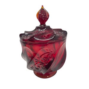 May include: A red glass jar with a lid and a decorative finial. The jar has a swirling design and ornate detailing. The finial is a flame-like shape with orange and yellow accents. The jar sits on a small, round base.