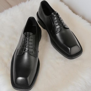 以下が含まれることがあります： 黒い革製のドレスシューズのペア。つま先は四角く、レースアップで留めます。靴は滑らかで光沢のある仕上がりで、厚いソールが付いています。靴は白くふわふわした表面に展示されています。