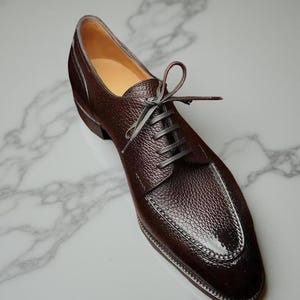 May include: A single brown leather dress shoe with a textured surface and gray laces. The shoe features a stitched design around the toe and a small heel. The shoe is displayed on a white marble surface.