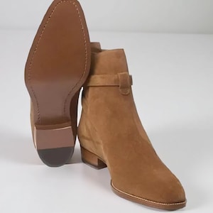 Op de afbeelding: Bruine suède enkellaarsjes met een lage hak en een bandje. De zool van één laars is zichtbaar, met de stiksels en constructie. De laarzen hebben een warme, neutrale kleur, geschikt voor diverse outfits. De hakhoogte is ongeveer 3 cm.