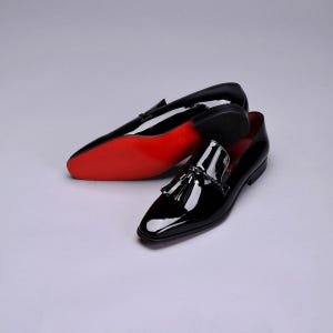 Può includere: Un paio di mocassini neri in vernice con dettagli a nappe. Le scarpe hanno una finitura lucida e suole rosse. I mocassini sono esposti su uno sfondo bianco.