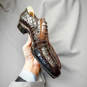 Può includere: Una scarpa da uomo in pelle marrone con una texture a pelle di coccodrillo. La scarpa ha una punta quadrata e un inserto in pelle marrone sulla tomaia. La scarpa è tenuta in mano.