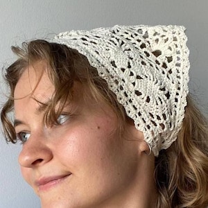 May include: Cream-colored, crocheted headscarf with a triangular shape. The head covering features an open, floral-like pattern. A delicate pearl necklace with a small pendant is also visible.