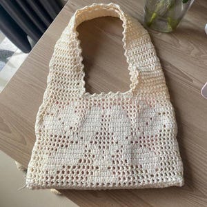 May include: Cream-colored crochet tote bag with a rectangular body and a scalloped edge. The bag features an openwork design and two handles. The bag is sitting on a wooden surface.