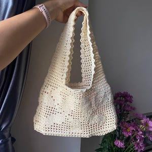 K&ouml;nnte beinhalten: Cremefarbene H&auml;keltasche mit gewelltem Rand. Die Tasche hat einen rechteckigen K&ouml;rper und zwei Henkel. Die Tasche wird von einer Hand gehalten. Im Hintergrund befindet sich ein Strau&szlig; lila Blumen.