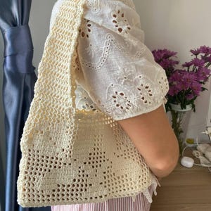 K&ouml;nnte beinhalten: Cremefarbene H&auml;keltasche mit floralem Muster. Die Tasche wird &uuml;ber der Schulter getragen. Die Person tr&auml;gt eine wei&szlig;e Bluse mit Lochstickerei und eine Hose mit rosa und wei&szlig;en Streifen.