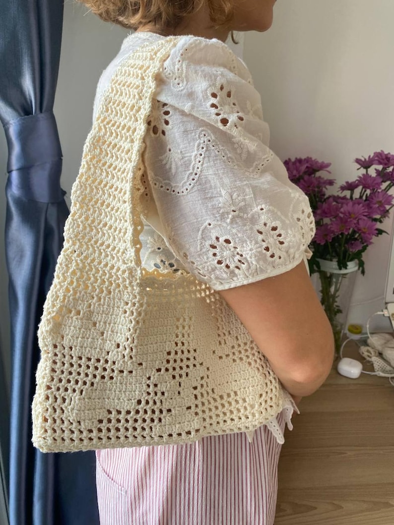 K&ouml;nnte beinhalten: Cremefarbene H&auml;keltasche mit floralem Muster. Die Tasche wird &uuml;ber der Schulter getragen. Die Person tr&auml;gt eine wei&szlig;e Bluse mit Lochstickerei und eine Hose mit rosa und wei&szlig;en Streifen.
