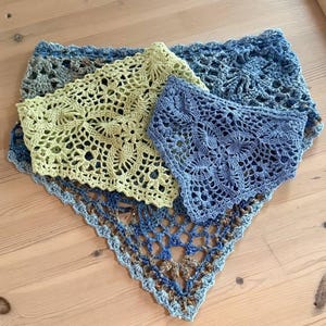 Op de afbeelding: Drie handgemaakte gehaakte sjaals in blauw- en groentinten zijn gerangschikt op een houten oppervlak. De sjaals hebben ingewikkelde open patronen, met een driehoekige vorm. Het kleurenpalet omvat lichtblauw, olijfgroen en donkerdere blauwtinten.